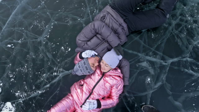 Young-couple-has-fun-during-winter-walk-against-background-of-ice-of-frozen-lake.-Lovers-lie-on-clear-ice-with-cracks,-have-fun,-kiss-and-hug.-View-from-above.-Honeymoon.-Love-story.