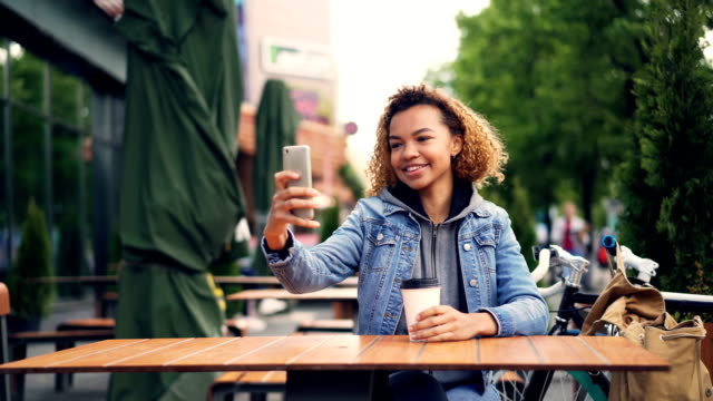 Unvorsichtige-afroamerikanische-Frau-froh-Tourist-nimmt-Selfie-mit-Smartphone-im-Straßencafé-mit-Take-Away-Kaffee-am-Tisch-sitzen,-Fahrrad-und-Rucksack-sind-sichtbar.