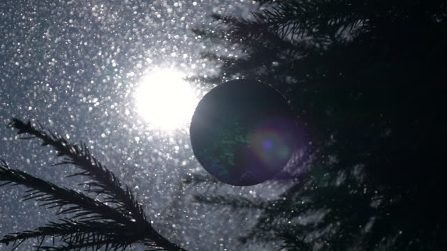 Winter-lyric-scene-with-sun-in-frame-with-lens-flare-and-green-and-blue-scaly-Christmas-tree-ball-in-back-sun-light,-waving-and-rotating-on-fir-branch-in-snowstorm-with-slow-motion.