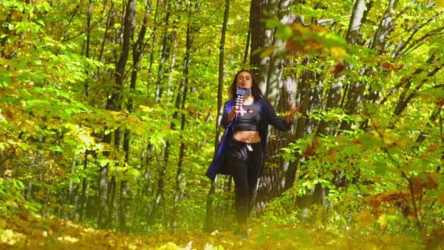 Blogging-charismatic-young-woman-is-walking-in-sunny-autumn-forest-and-recording-video-for-vlog-using-camera-slow-motion