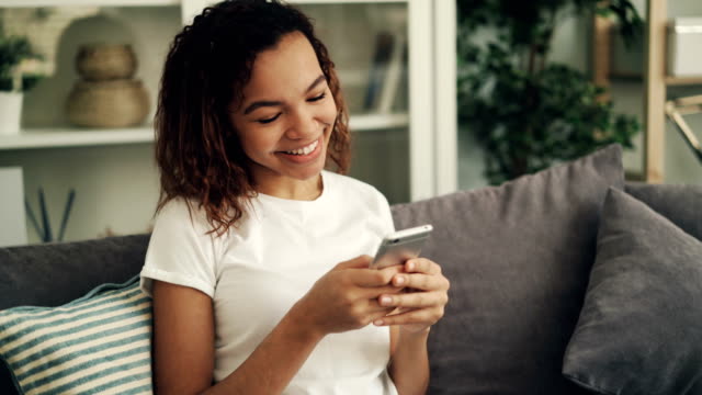 Fröhliche-Afroamerikanerin-hält-Smartphone,-berührt-Bildschirm-und-lacht-auf-Couch-im-hellen-Raum.-Junge-Leute,-Gadgets-und-Spaß-Konzept.