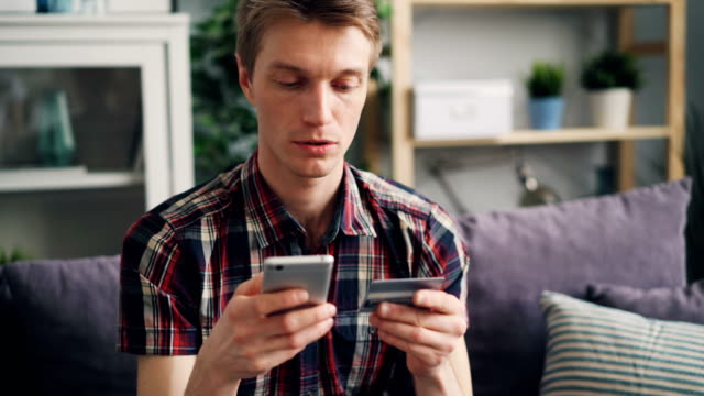 Junger-Mann-macht-Online-Zahlung-mit-Bankkarte-mit-Smartphone-zu-Hause