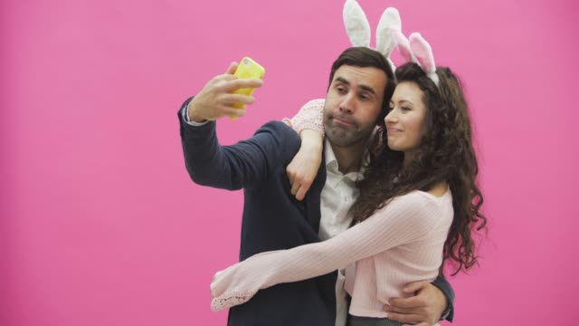 Young-tender-pair-standing-on-a-pink-background.-Looking-at-the-phone-while-doing-selfie-on-the-mobile-phone-with-the-ears-of-a-pink-rabbit-on-the-head.-Happy-family-is-preparing-for-Easter