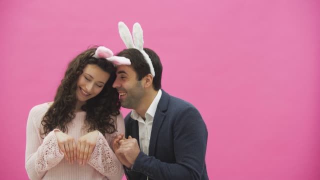 Beautiful-young-couple-standing-on-pink-background.-During-this-time,-they-recreate-the-movements-of-hares,-placing-their-hands-like-a-rabbit.-Together-with-pink-barking-ears-on-the-head.-Happy-family-is-preparing-for-Easter.