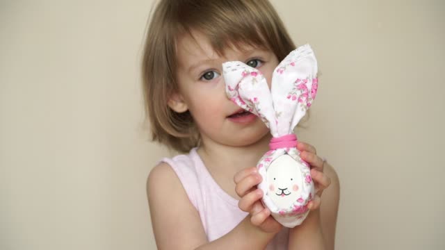 Portrait-of-little-pretty-smiling-caucasian-girl-holds-in-hand-chicken-egg-decorated-for-Easter-bunny,-with-painted-muzzle-and-ears.