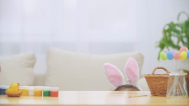 Playful-little-girl-with-bunny-ears-is-hiding-under-the-table-full-of-Easter-decorations.-Nice-cute-white-bunny-is-jumpimg-into-and-attacking-girl's-head-in-a-playful-form.-Catching-game.