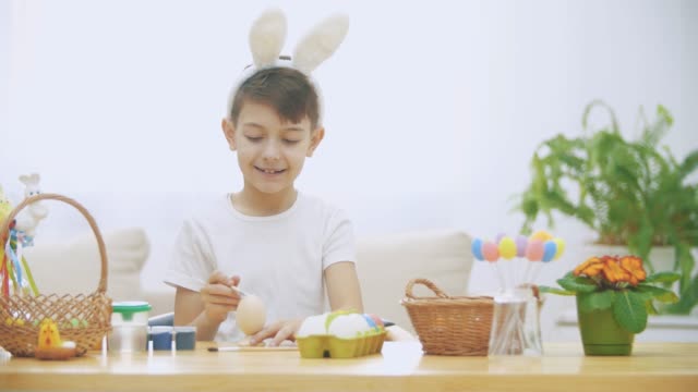 Little-cute-boy-is-having-fun-holding-a-paint-brush-in-her-right-hand.-Boy-is-watching-at-nude-paint-brushes-and-is-trying-to-colour-the-Easter-egg,-sitting-at-the-wooden-table-with-Easter-decorations.