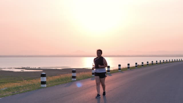 Übergewichtige-asiatische-Frauen-joggen-am-frühen-Morgen-im-Sonnenlicht-auf-der-Straße.-Konzept-des-Gewichtsverlustes-mit-Bewegung-für-die-Gesundheit.-zeitlupe