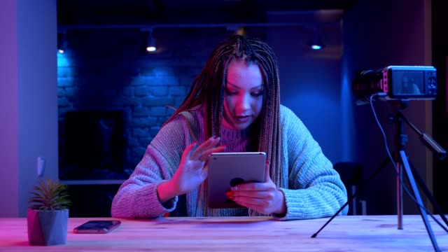 Closeup-shoot-of-young-attractive-female-blogger-with-dreadlocks-texting-on-the-tablet-smiling-cheerfully-streaming-live-with-the-neon-background-indoors