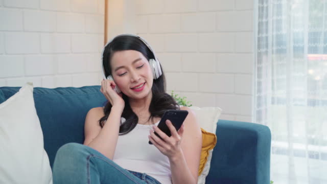 Asiatische-Frau,-die-Musik-hört-und-mit-dem-Smartphone,-weiblich-mit-Entspannungszeit-auf-dem-Schlafsofa-im-Wohnzimmer-zu-Hause.-Fröhliche-weibliche-Hörmusik-mit-Kopfhörer-Konzept.