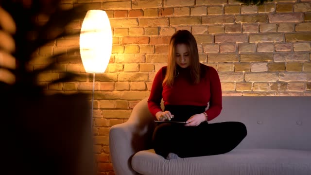 Nahaufnahme-von-jungen-hübschen-kaukasischen-weiblichen-TEXTen-auf-dem-Tablet-dann-Blick-auf-die-Kamera-mit-Nachdenklichkeit-sitzen-auf-der-Couch-drinnen-sitzen