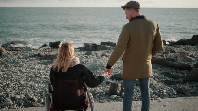 Loving-pair-is-standing-on-seacoast,-disabled-woman-and-healthy-man,-back-view