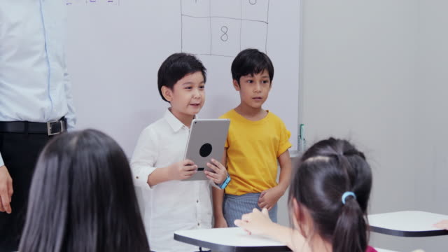 Niño-explicando-el-tema-al-estudiante-en-el-salón-de-clases.-Niño-usando-tableta-para-ayudar-en-su-tema.-Concepto-de-Educación-y-Tecnología.
