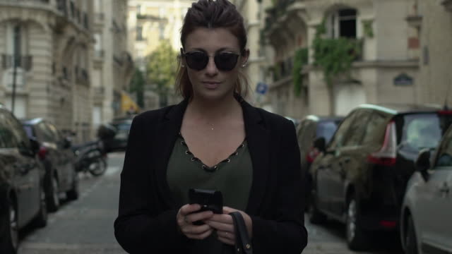 Attractive-caucasian-classy-woman-with-sunglasses,-freckles,-piercings,-black-jacket-and-red-hair-writing-a-text-message-smiling-walking-through-the-street,-during-sunny-day-in-Paris.-Slow-motion.