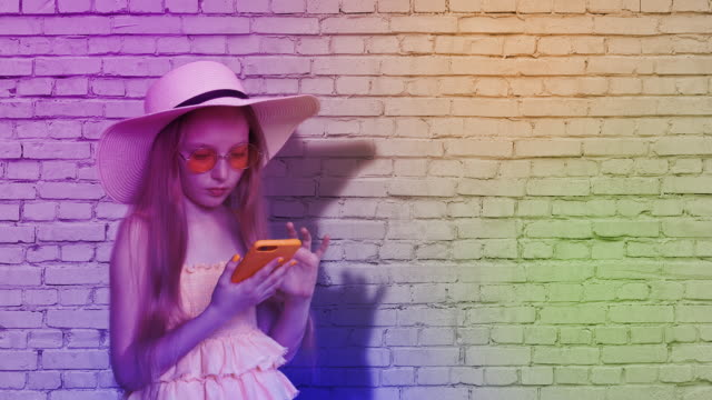 Teenager-girl-browsing-mobile-phone-on-brick-wall-background-in-colorful-light.-Young-girl-using-smartphone-for-surfing-in-social-networks-on-multicolored-brick-wall-background.