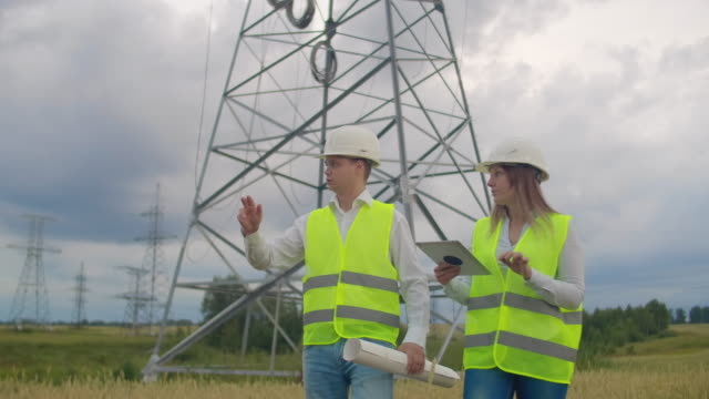 Power-lines,-man-and-woman-engineers-with-a-tablet-in-their-hands-check-the-progress-of-the-installation-of-new-towers-and-analyze-the-network.