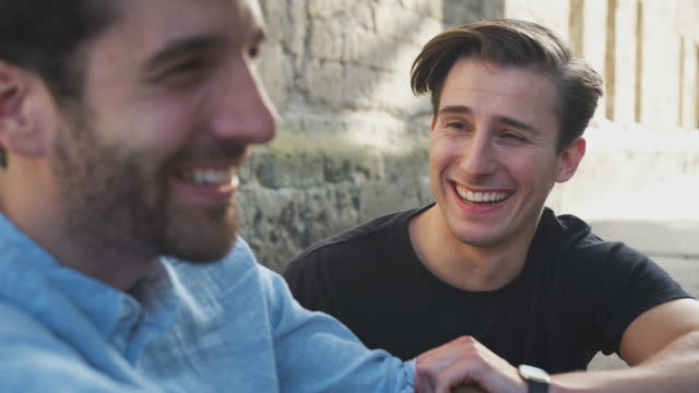 Male-Gay-Couple-Sitting-Outdoors-On-Steps-Of-Building-Laughing-Together