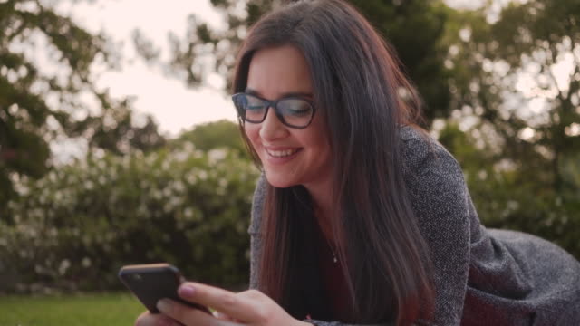 Porträt-einer-attraktivlächelnden-jungen-Frau,-die-im-Park-liegt-und-SMS-Nachrichten-auf-dem-Smartphone-abgibt