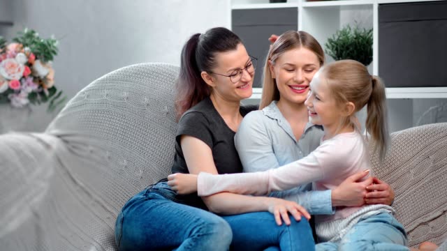 Same-sex-female-family-smiling-hugging-daughter-sitting-on-couch-looking-at-camera