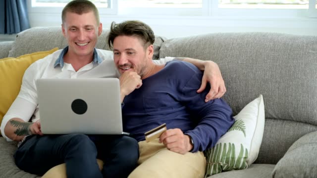 Gay-couple-relaxing-on-couch-using-laptop-computer.-Talking-and-holding-card.
