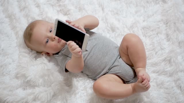 Lindo-bebé-sostiene-el-teléfono-en-las-manos.-El-niño-juega-con-el-teléfono-celular,-lo-examina,-tira-en-sus-manos-y-trata-de-comer-teléfono-inteligente-poniéndolo-en-su-boca