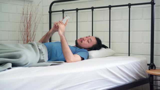 Junge-asiatische-Mann-aufwachen-am-Morgen,-Asia-männlich-mit-Handy-überprüfen-Social-Media-und-senden-Nachricht-nach-wach-auf-dem-Bett-im-Schlafzimmer-zu-Hause.-Schöne-Männer-nicken,-verschlafen-entspannen-in-modernen-HausKonzept.