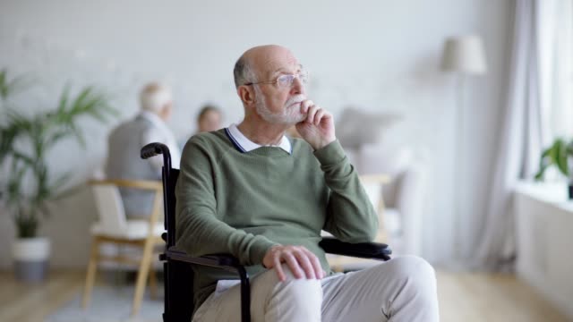Tracking-medium-shot-of-disabled-senior-man-in-eyeglasses-sitting-in-wheelchair-looking-away-and-thinking-in-assisted-living-home