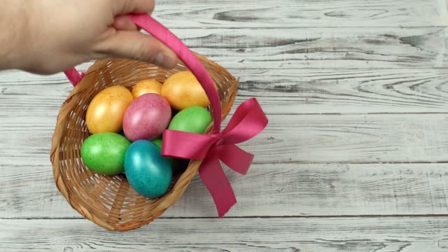 Male-hand-puts-a-gift-basket-with-colorful-chicken-eggs-on-a-wooden-white-background,-Easter-holiday,-copy-space,-celebration