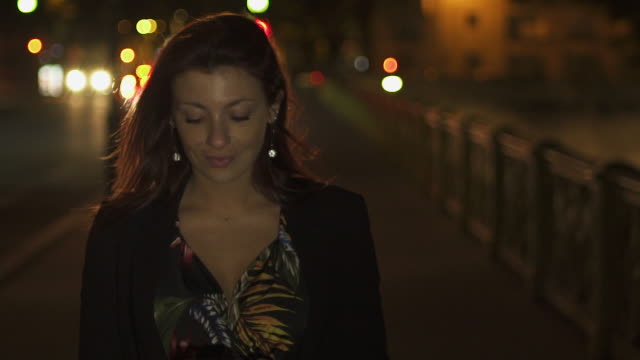 Cute-Happy-caucasian-modern-woman-wearing-flower-dress,-black-jacket-and-red-hair-walking-through-the-street-and-writing-a-text-message-on-her-smartphone-by-night.-Paris-4K-UHD.-Slow-Motion.