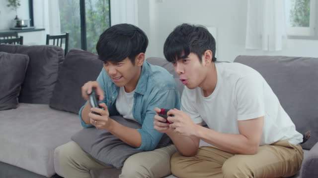 Young-Asian-gay-couple-play-games-at-home,-Teen-korean-LGBTQ-men-using-joystick-having-funny-happy-moment-together-on-sofa-in-living-room-at-house.-Slow-motion-shot.