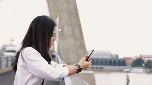 Porträt-von-attraktiven-jungen-asiatischen-Frau-mit-Smartphone-Chat-mit-Freunden-und-Surfen-in-sozialen-Medien-auf-einem-Handy,-während-auf-der-Brücke-stehen.
