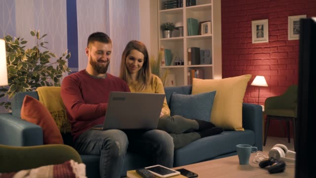 Feliz-pareja-sentada-en-el-sofá-y-conectando-con-su-computadora-portátil