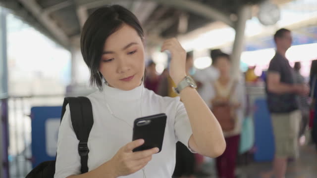 Retrato-de-mujer-asiática-con-auriculares-escuchando-música-y-usando-teléfono-inteligente-para-chatear-con-amigos-o-navegar-mientras-espera-un-tren.-Tecnología-en-la-vida-cotidiana-y-los-viajes.