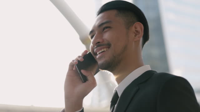 Hombre-de-negocios-asiático-con-un-traje-negro-usando-su-teléfono-inteligente-hablando-por-teléfono-mientras-estaba-de-pie-en-la-calle-cerca-de-un-gran-edificio-de-oficinas-urbano-con-luz-solar.