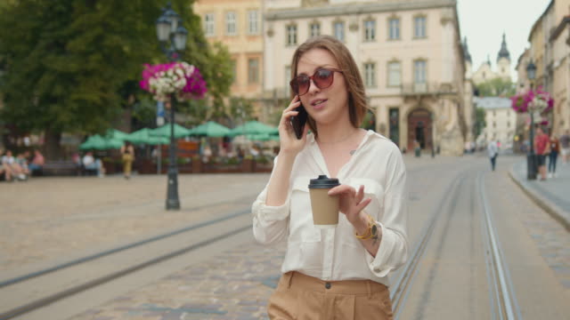 The-girl-is-talking-on-the-phone.-She-is-walking-and-smiling.-She's-holding-a-cup-of-coffee.-She's-wearing-sunglasses.-The-people-are-walking-in-the-background.-4K