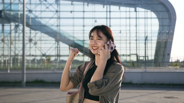 Appealing-cheerful-asian-young-woman-talking-on-smartphone-on-the-background-of-airport-building