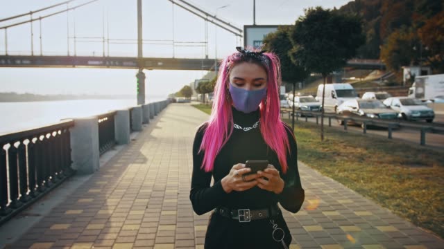 Hipster-female-with-pink-hair,-in-informal-outfit-and-protective-mask-is-typing-on-mobile-phone,-walking-by-embankment