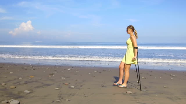 CLOSE-UP-Woman-with-crutches-walking-along-the-splashing-ocean-on-sunny-Bali