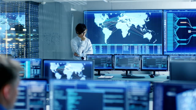 In-the-System-Control-Room-Chief-Engineer-Thinks-While-Standing-Before-Big-Screen-with-Interactive-Map-on-it.-Data-Center-is-Full-of-Monitors-Showing-Graphics.