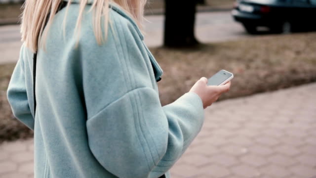 Slow-motion-blonde-teen-in-blue-coat-texting.-Side-view.-Girl-with-long-hair-reading-from-smartphone.-Web-streaming
