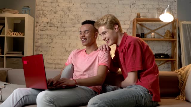 Two-multinational-homosexual-friends-sit-on-the-couch,-use-a-laptop,-watch-a-funny-video-on-the-Internet.-Home-cosiness,-family,-internet-concept.-60-fps