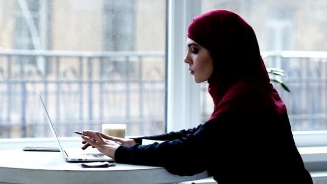 Indoors-footage-of-beautiful-muslim-girl-with-hijab-on-her-head-typing-something-and-then-writing-something-down-in-her-notebook