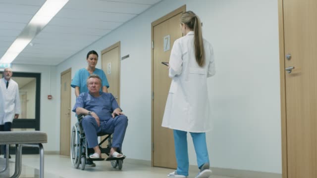 Arzt-spricht-mit-ihnen-zwar-im-Krankenhaus-Flur,-Krankenschwester-schiebt-älteren-Patienten-im-Rollstuhl,-Tablet-Computer-verwenden.-Sauberes,-neues-Krankenhaus-mit-medizinischen-Fachpersonal.