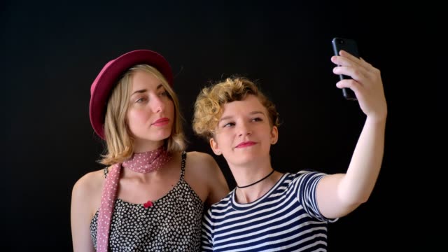 Zwei-hübsche-junge-blonde-Frauen-umarmen-und-nehmen-Selfie,-isoliert-auf-schwarz-Studio-Hintergrund
