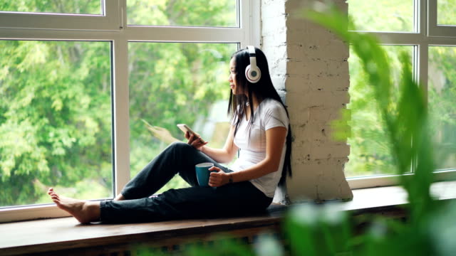 Pretty-Asian-student-is-enjoying-music-in-headphones-sitting-on-window-sill-and-using-smartphone-touching-screen.-Modern-lifestyle-and-technology-concept.
