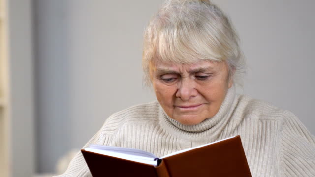 Aged-lady-in-wheelchair-holding-book-and-looking-for-eyeglasses-lying-on-table