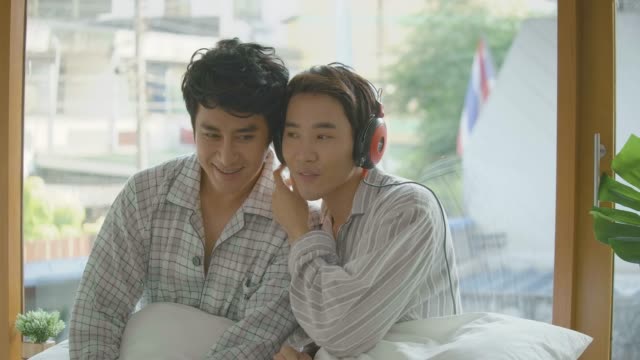 Junge-asiatische-Schwule-paar-sitzt-auf-der-Fensterbank-Buch-lesen-und-hören-von-Musik-zu-Hause-am-Morgen.