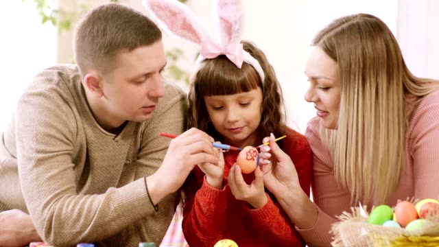 the-girl-holds-an-Easter-egg,-and-dad-and-mom-decorate-it.