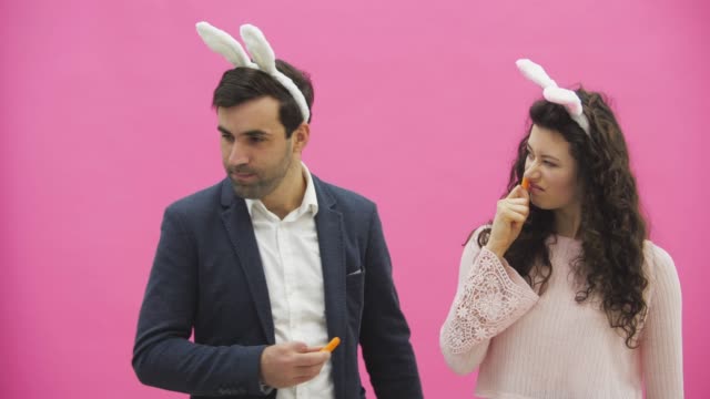 Young-couple-are-beautiful-on-pink-background.-During-this-time,-they-are-dressed-in-rabble-ears.-Looking-at-each-other,-they-play-carrots,-showing-different-rabbit-movements.
