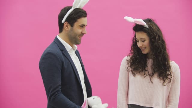 Young-creative-couple-on-pink-background.-With-hackneyed-ears-on-the-head.-During-this-man-gives-a-soft-toy-with-a-hare-to-his-wife.-Embracing-a-girl-looking-into-the-camera-and-showing-a-gesture-class.